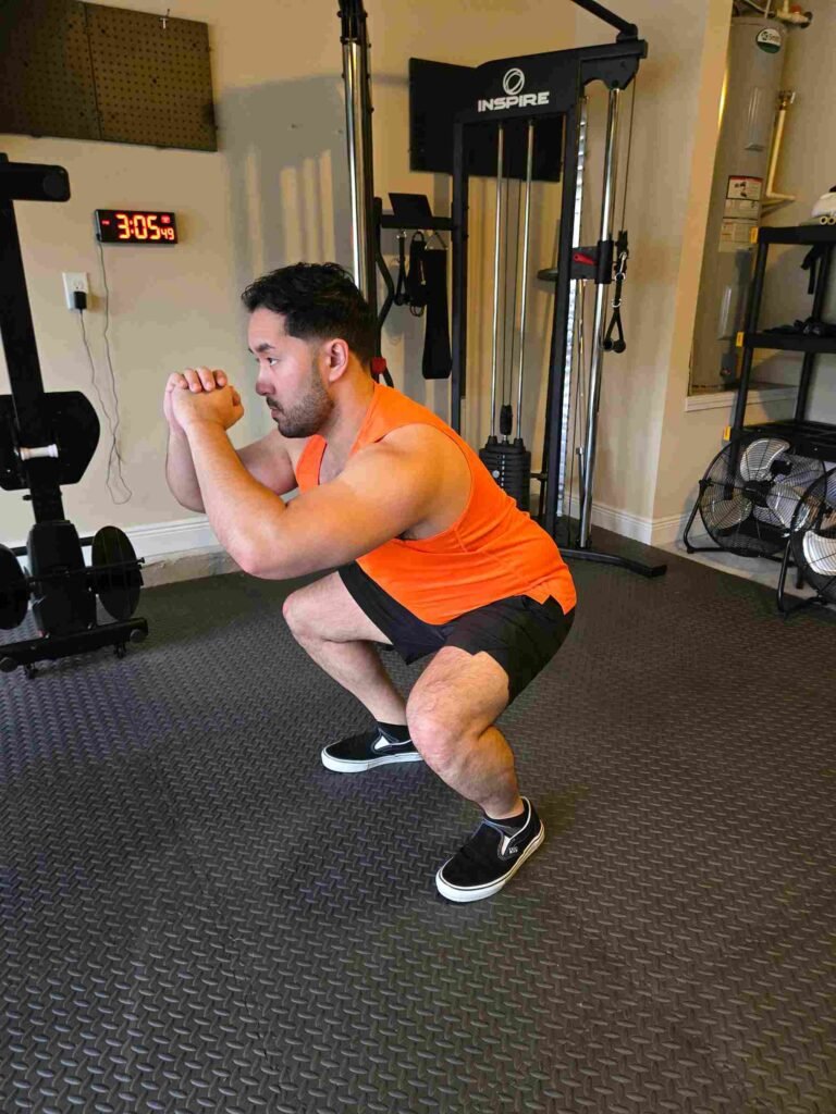 Bodyweight Squat in Home Gym A man performing a bodyweight squat in a home gym, wearing an orange tank top and black shorts, focusing on lower body strength and mobility.