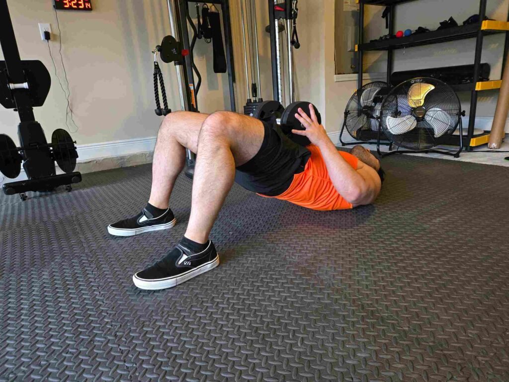 Dumbbell Glute Bridge in Home Gym A man performing a glute bridge with a dumbbell on his hips in a home gym, wearing an orange tank top and black shorts, focusing on glute and lower body strength.