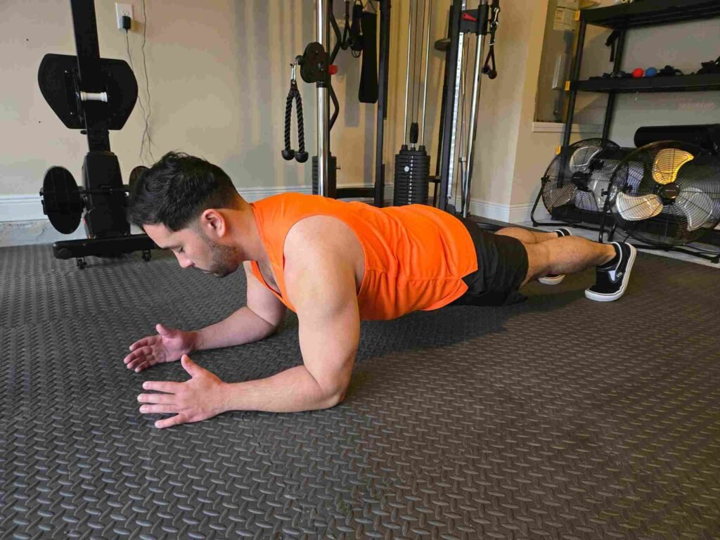 A man performing a forearm plank in a home gym, wearing an orange tank top and black shorts, focusing on core strength.