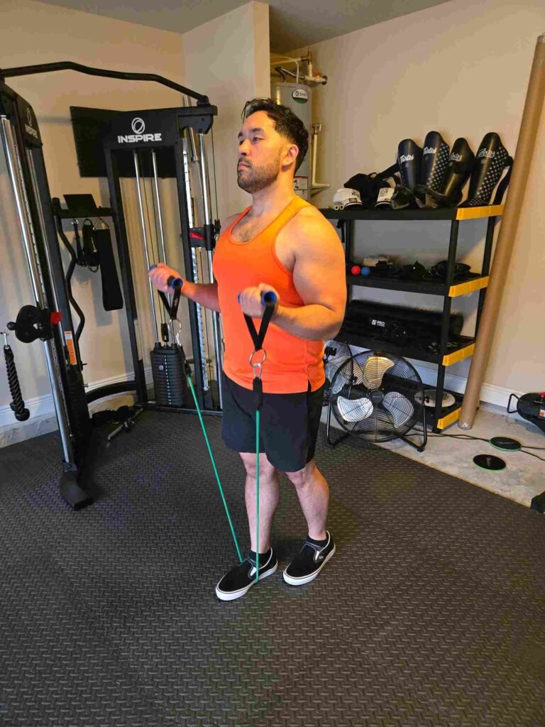 Resistance Band Bicep Curl in Home Gym A man performing a resistance band bicep curl in a home gym, wearing an orange tank top and black shorts, focusing on arm strength.
