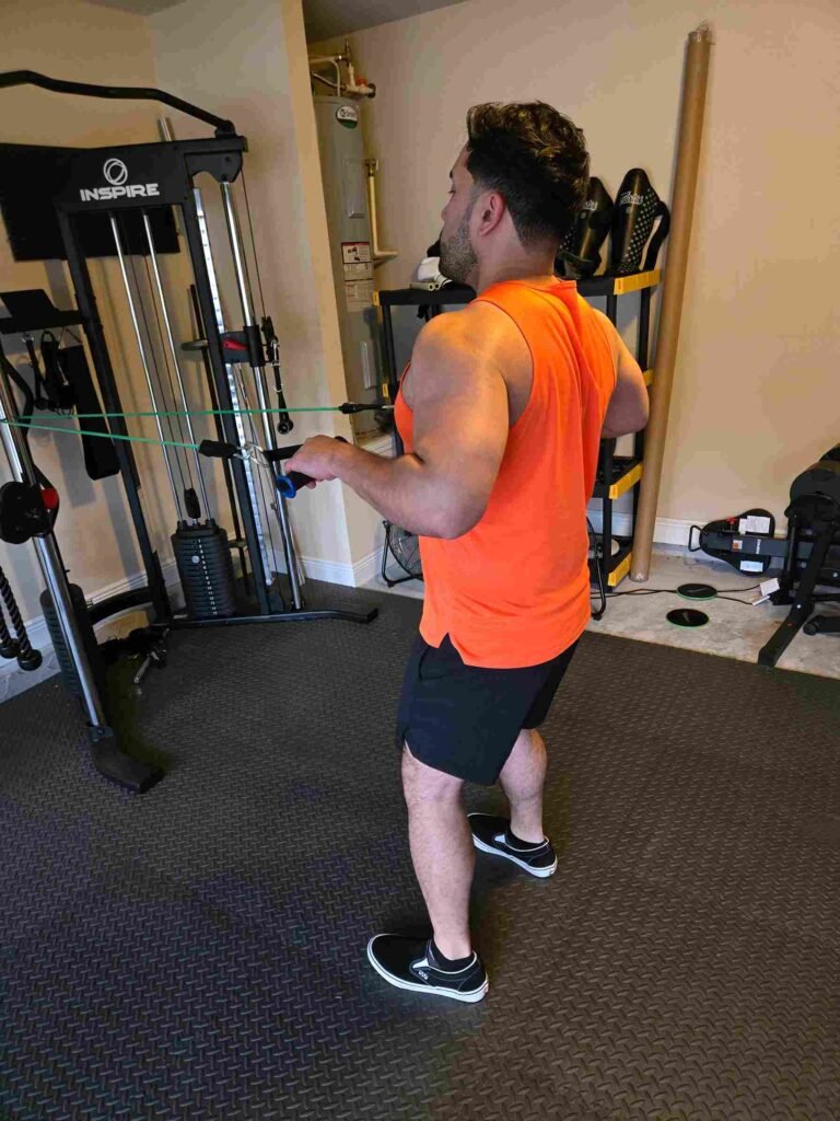 Resistance Band Row in Home Gym A man performing a resistance band row in a home gym, wearing an orange tank top and black shorts, with gym equipment visible in the background.