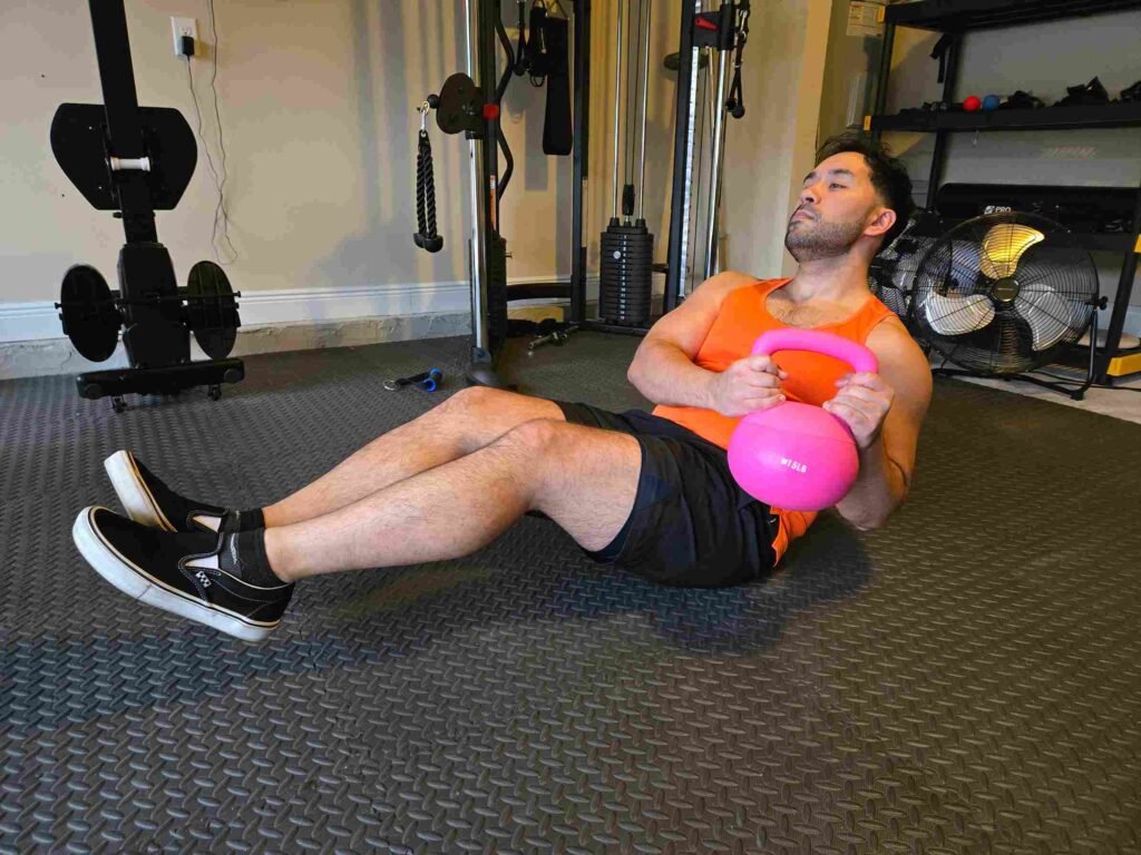 Russian Twist with Kettlebell in Home Gym A man performing a Russian twist with a pink kettlebell in a home gym, wearing an orange tank top and black shorts, focusing on core strength.